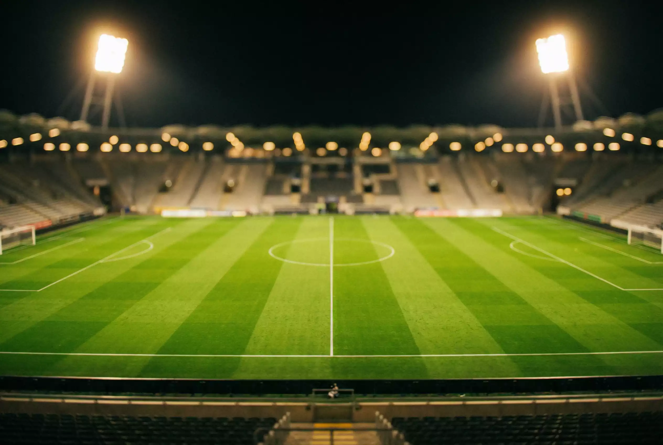 Campo de futebol visto de uma bancada com relvado verde e linhas brancas sob iluminação noturna