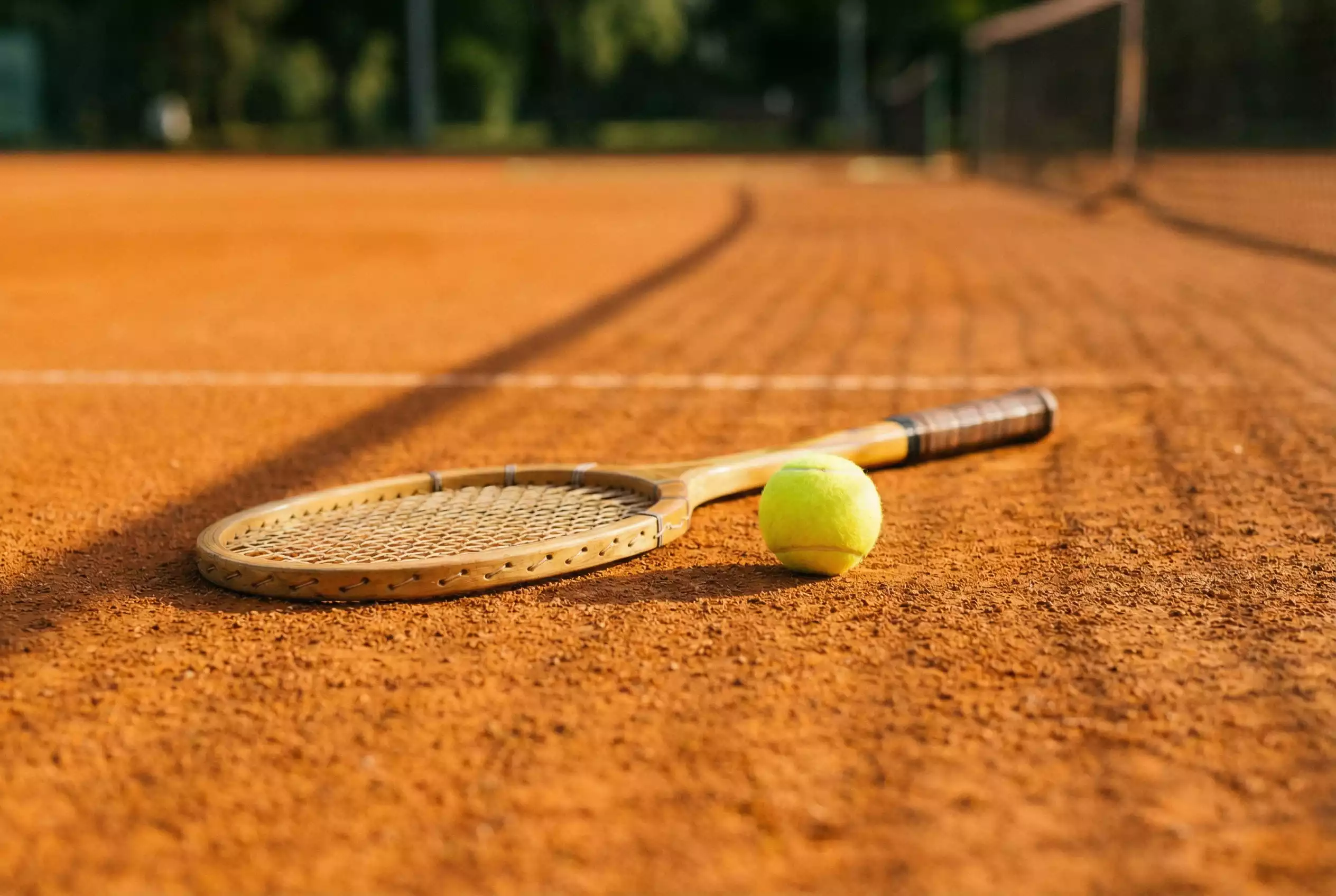 Raquete de ténis e bola amarela sobre um court de terra batida ao sol